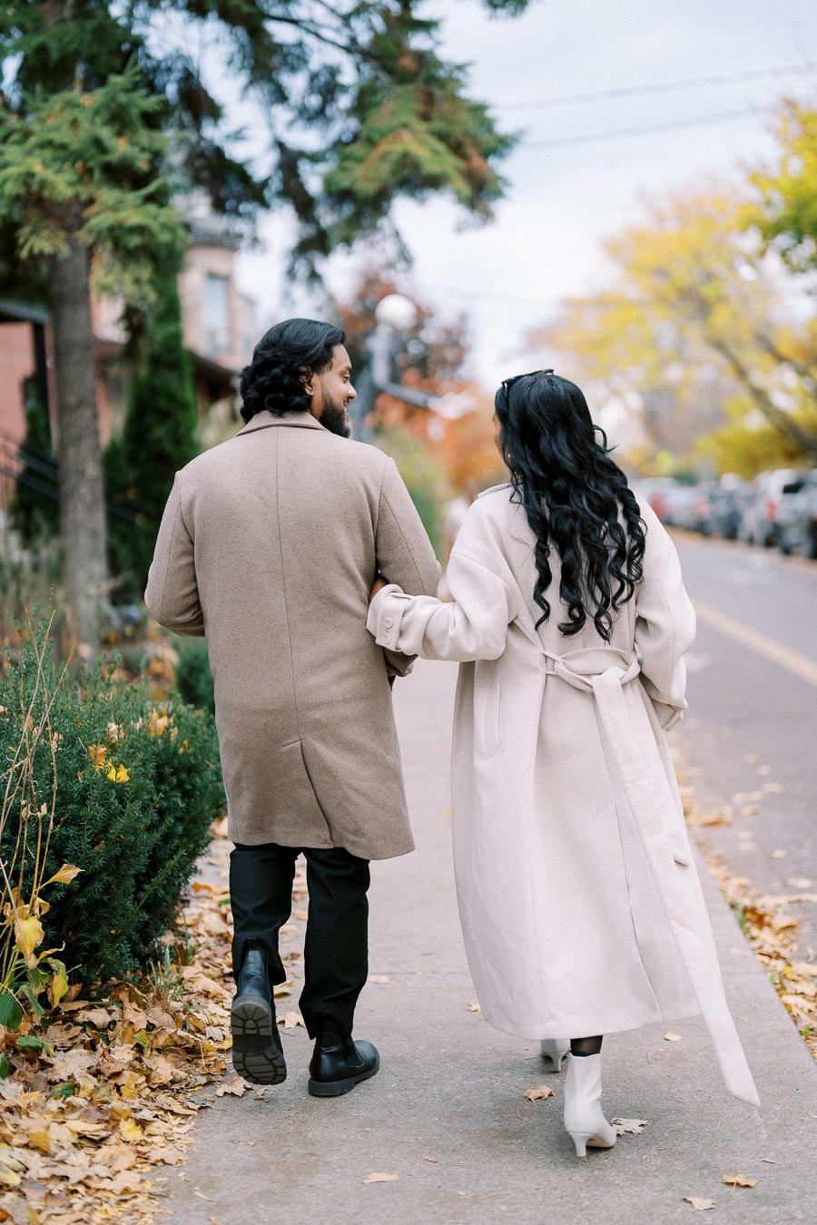 A couple walking arm-in-arm down a tree-lined sidewalk in autumn, wearing stylish long coats.