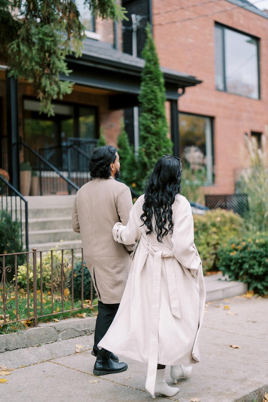 A couple in stylish coats walking hand in hand on a suburban street in autumn, with a brick house and greenery in the background.