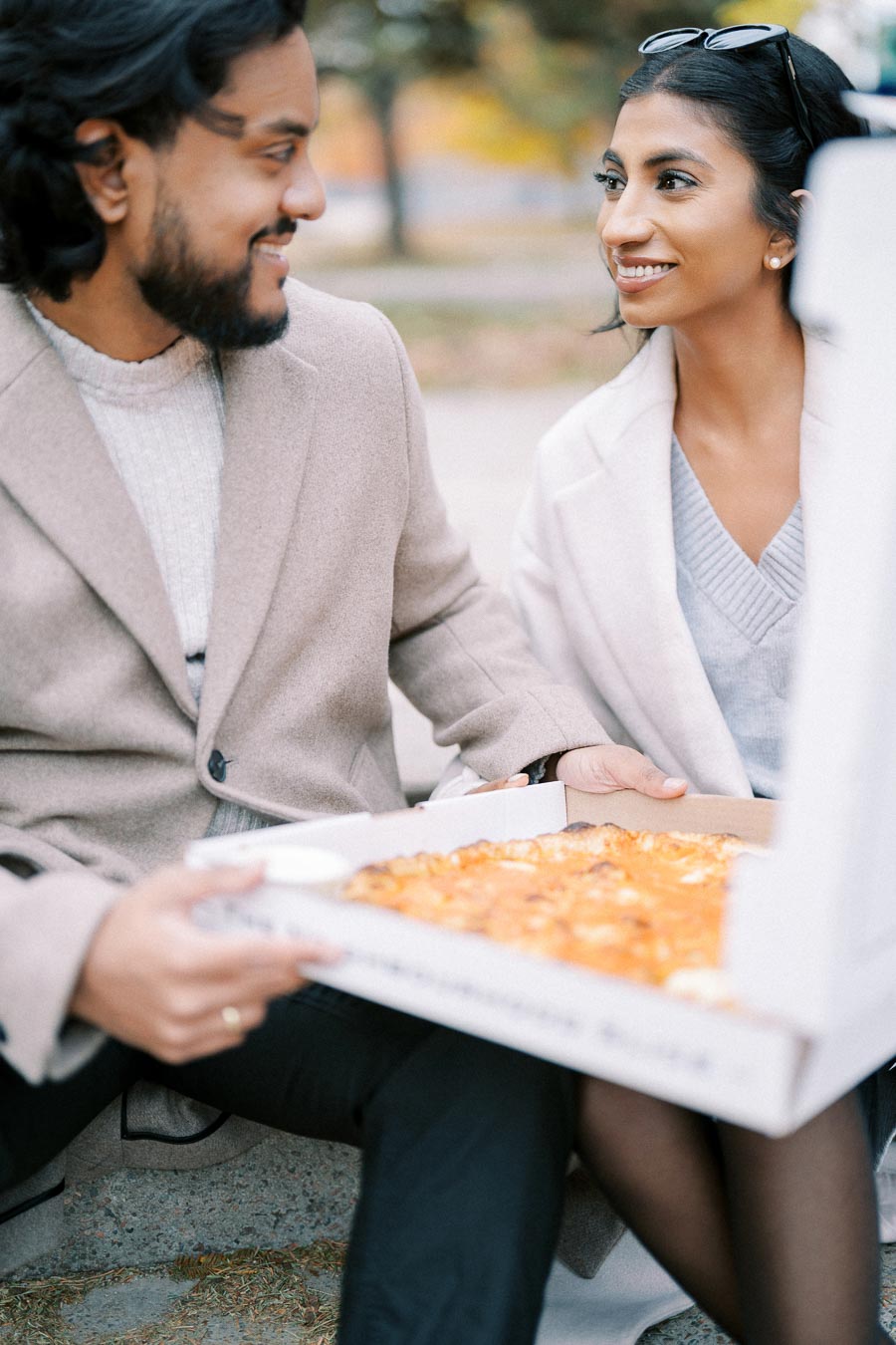 A couple enjoying a pizza together outdoors, sitting on a bench and dressed in cozy fall clothing, smiles and turns to each other.