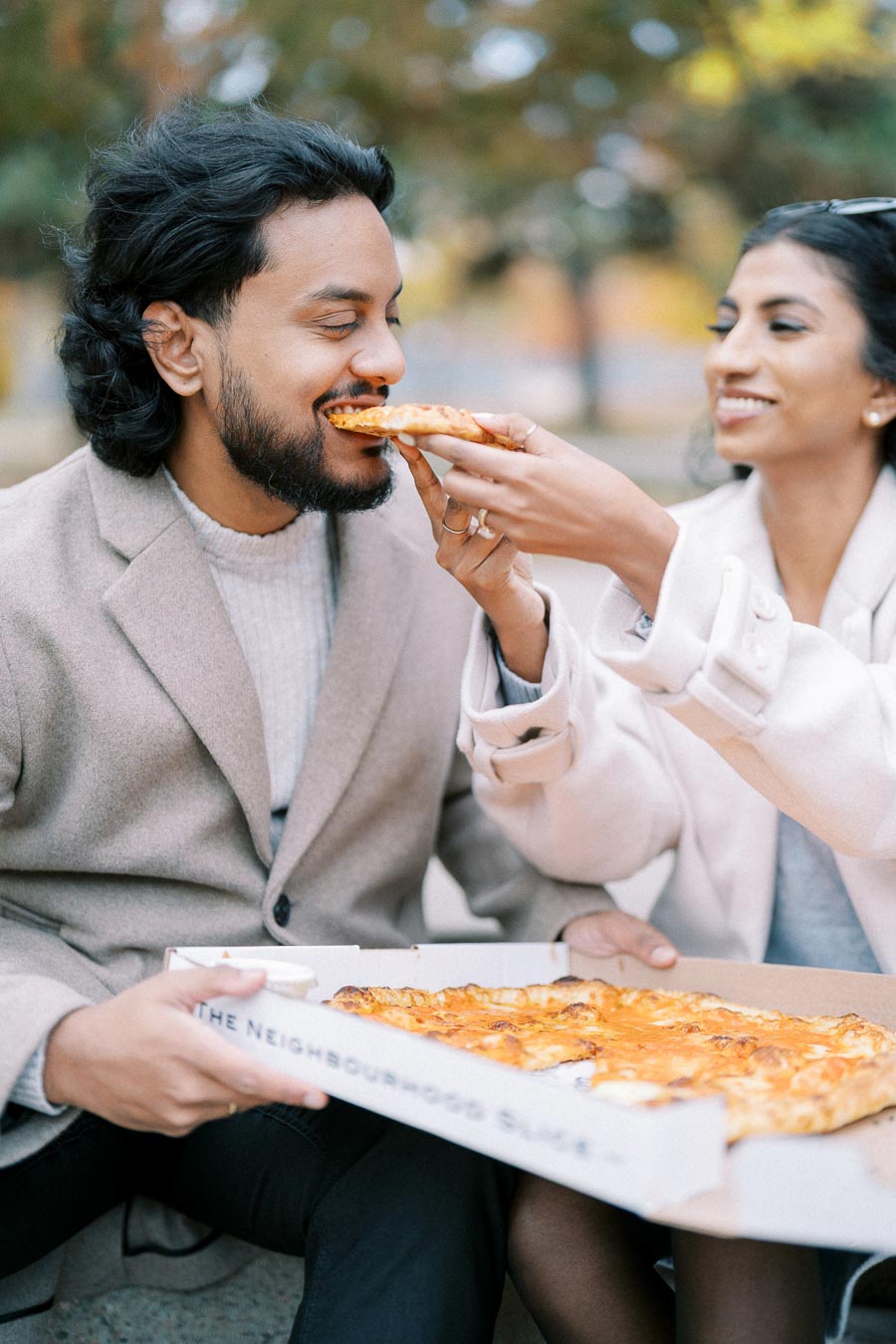 Couple enjoying a casual outdoor pizza date, with a woman playfully feeding a slice to her partner from an open pizza box, showcasing a joyful and relaxed moment.