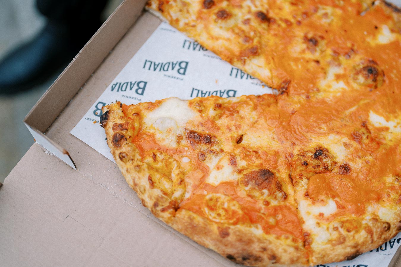 Close-up of a cheese pizza with a crispy, golden crust and melted cheese, served in a cardboard box lined with branded parchment paper.