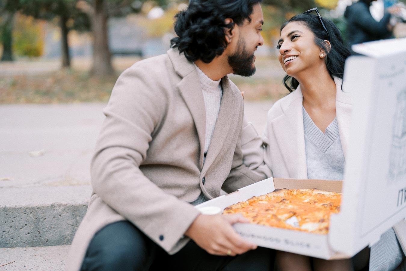 Couple sharing a romantic moment while enjoying pizza outdoors in a park setting, dressed warmly for fall.