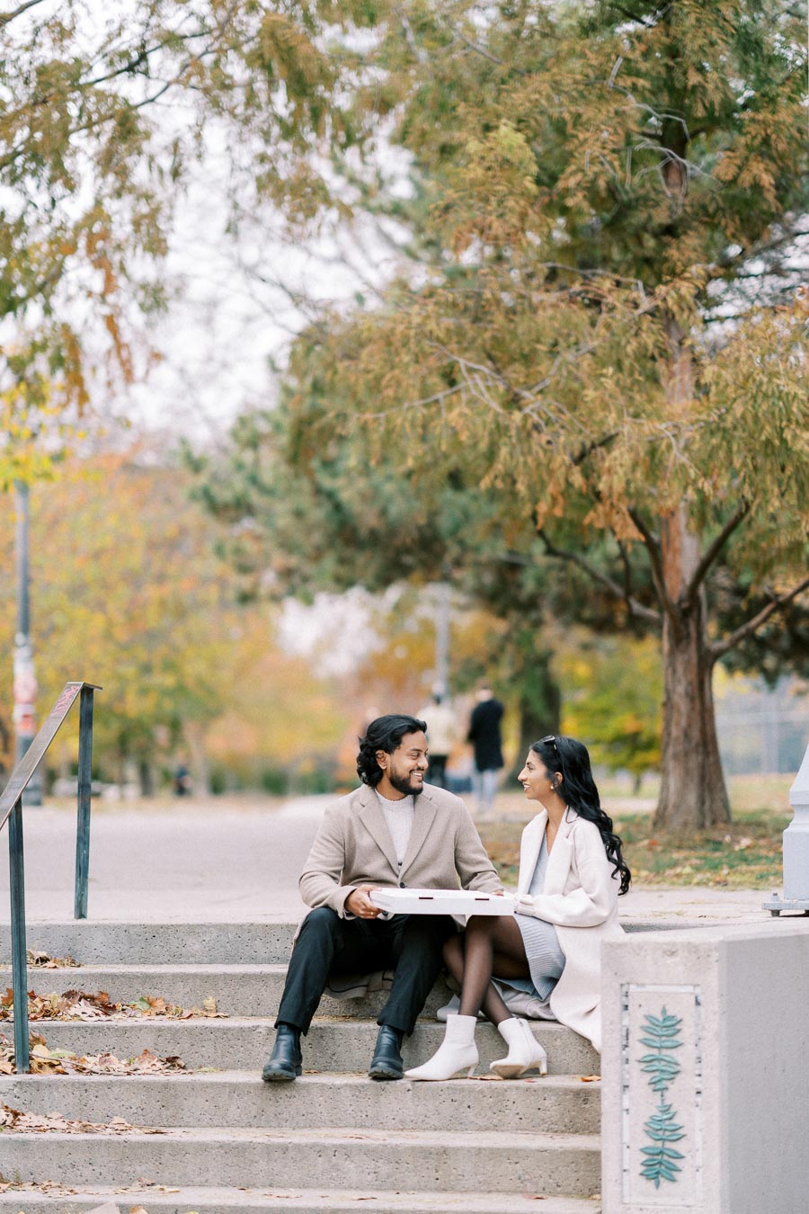 Couple sitting on park steps sharing a pizza on a fall day, surrounded by autumn trees and fallen leaves.