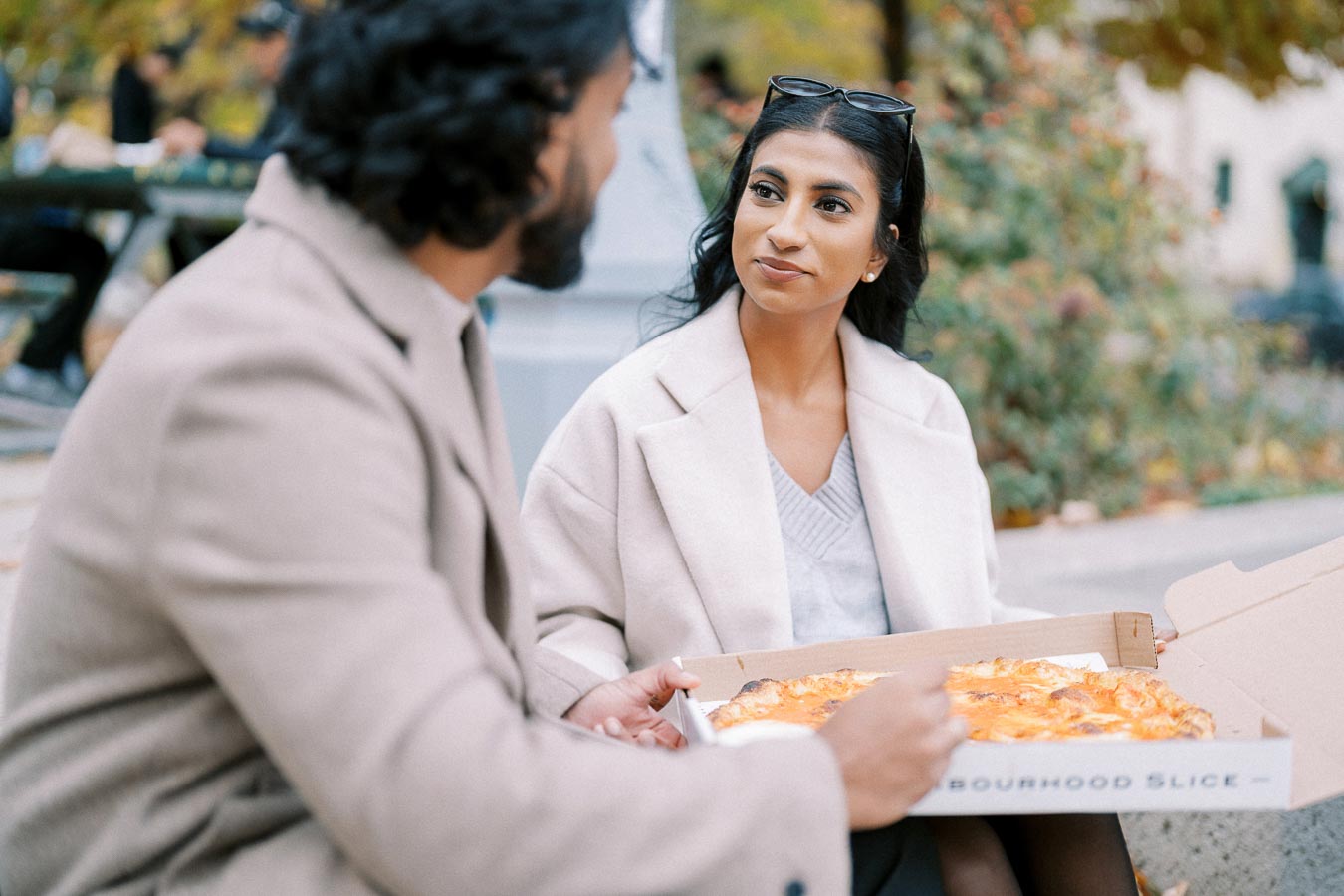 A man and woman sitting in a park enjoying slices of pizza from a box on a fall day, wearing beige coats and smiling at each other.