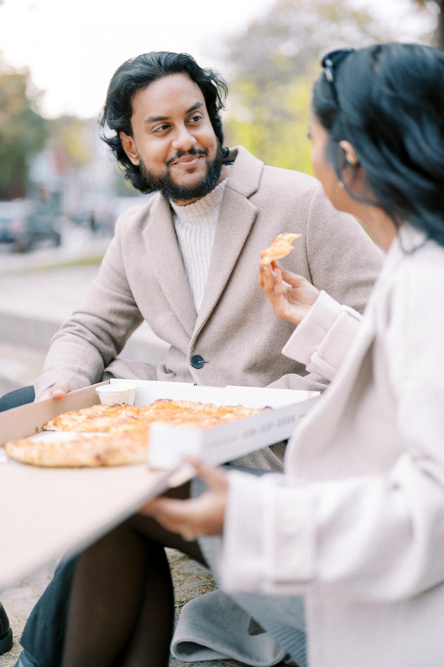 A couple in stylish coats enjoying pizza outdoors on a crisp fall day.