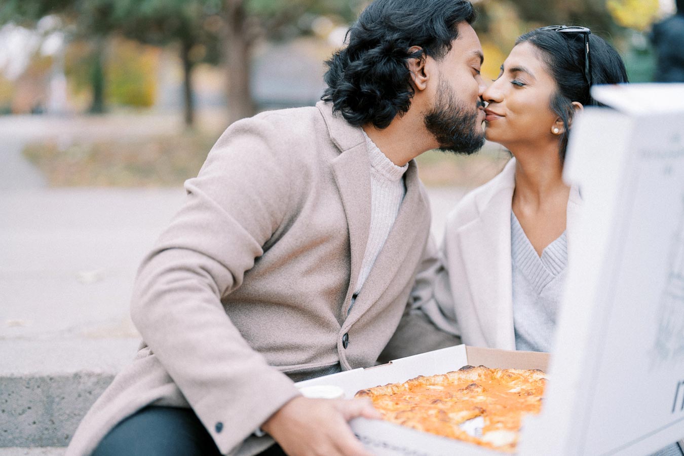 A couple sharing a romantic moment while holding a pizza box outdoors in a park setting, wearing matching beige coats and smiling at each other lovingly.