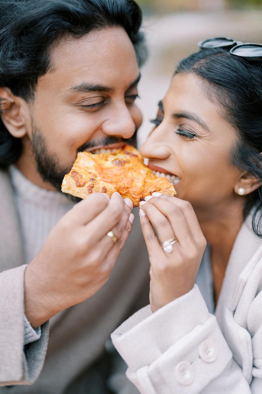 A happy couple sharing a slice of pizza, smiling and enjoying an outdoor moment together, dressed in cozy coats.