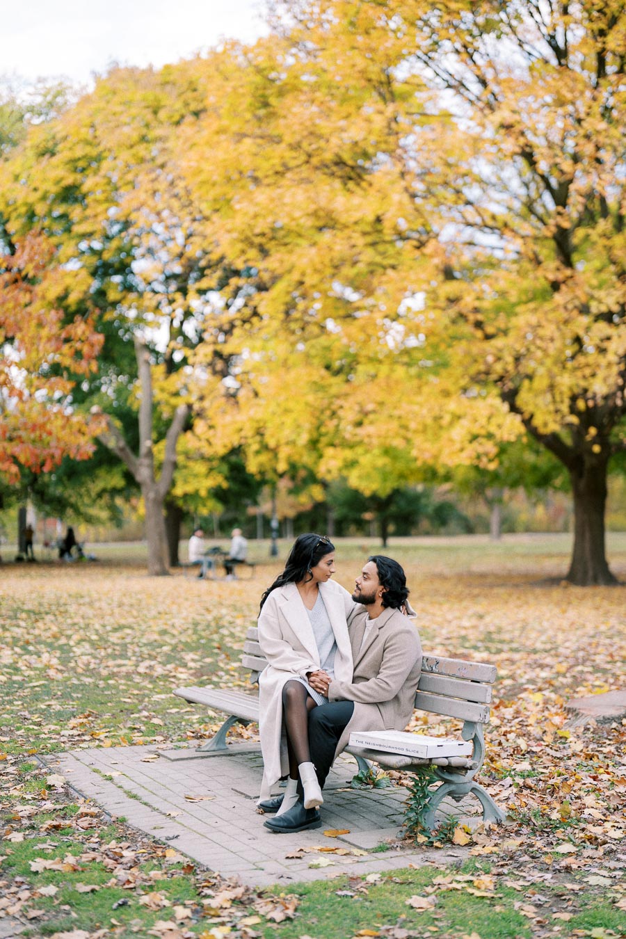 A couple sitting on a park bench surrounded by autumn foliage, with yellow and orange leaves covering the ground, creating a romantic and serene atmosphere.
