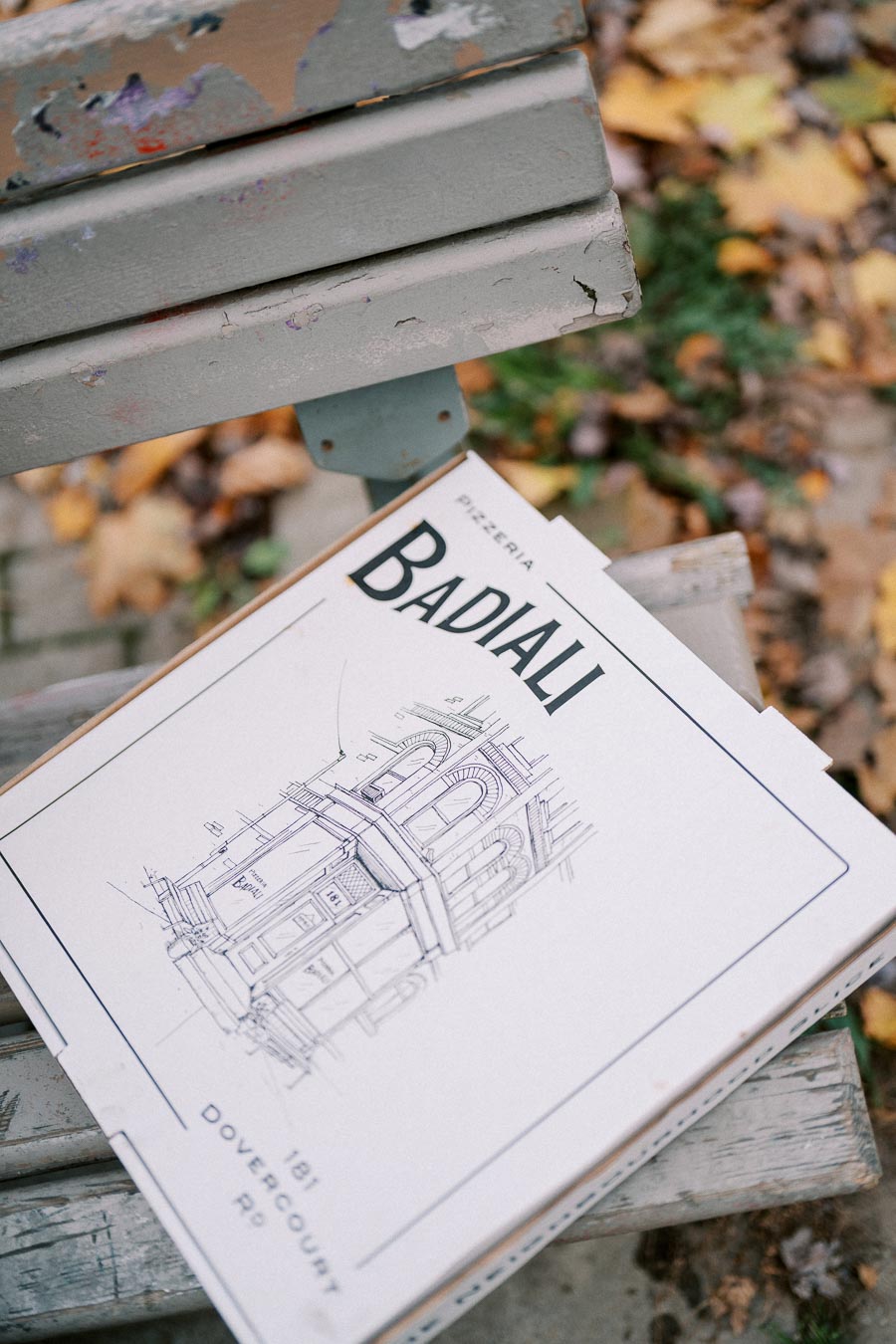 Pizza box from Pizzeria Badiali on a weathered park bench surrounded by autumn leaves, featuring an architectural drawing on the lid and the address 181 Dovercourt Rd.