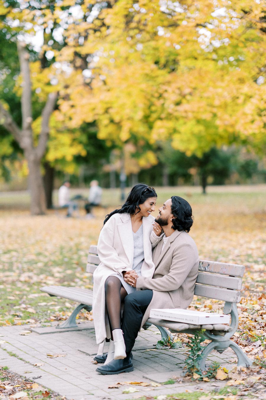 Couple sitting on a park bench during autumn, surrounded by colorful leaves with a backdrop of trees.