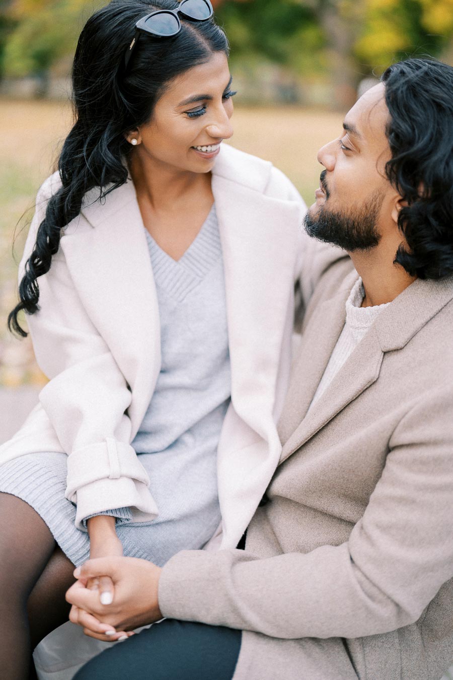 A smiling couple seated outdoors, wearing stylish neutral-toned coats, enjoying a moment of connection in a park setting.