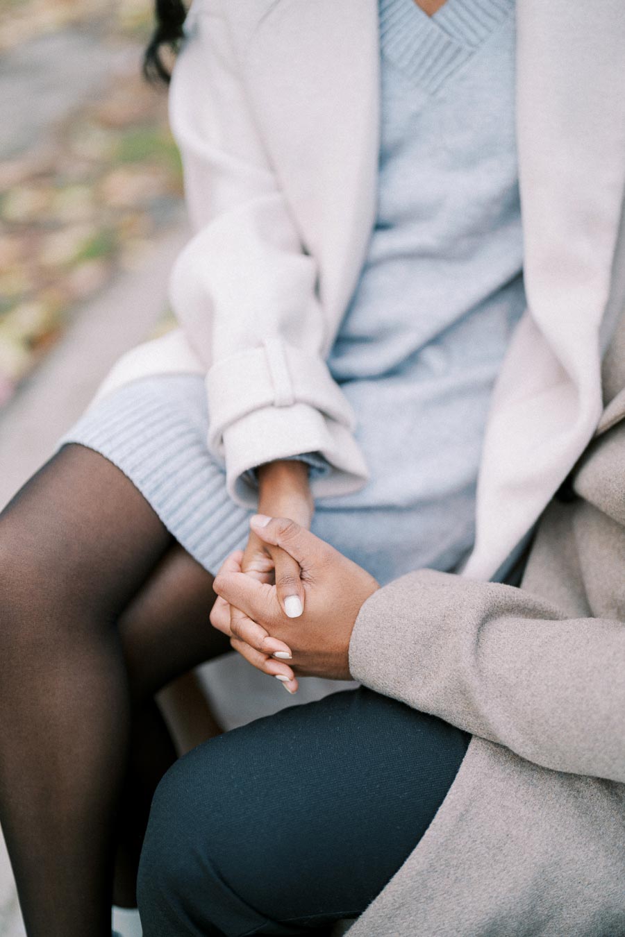 A close-up of a person wearing a stylish beige coat over a light blue dress sitting outdoors, with hands clasped and legs crossed on a bench with autumn leaves in the background.