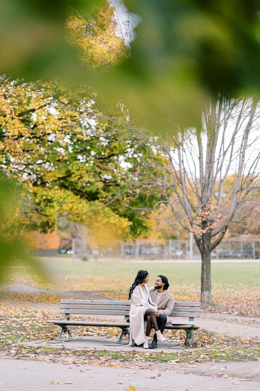 A couple sitting on a park bench surrounded by autumn leaves, engaging in a cozy conversation under a tree.