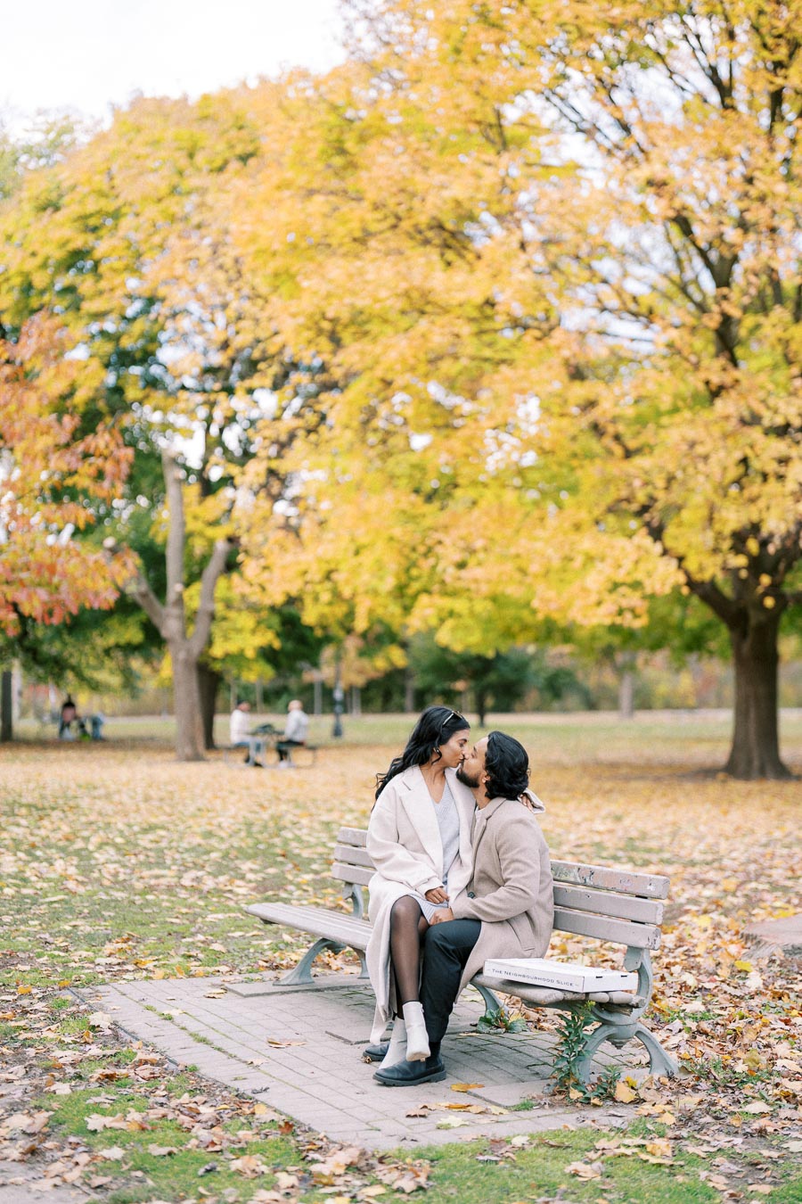 A couple sitting closely on a park bench surrounded by vibrant autumn foliage, with fallen leaves scattered on the ground, creating a romantic and serene atmosphere.