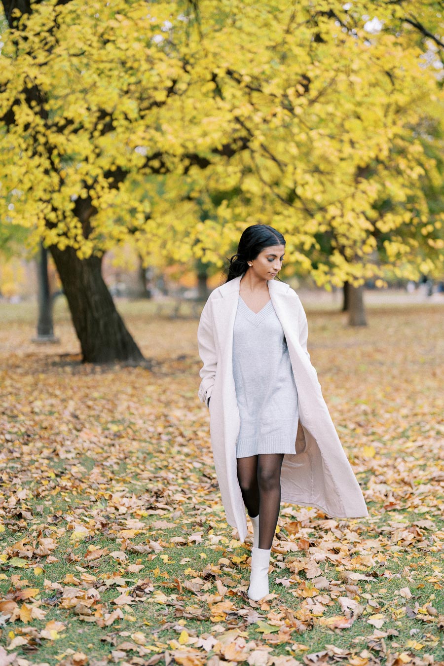 Woman in a white coat and dress walking through autumn leaves in a park, with yellow foliage on trees in the background.