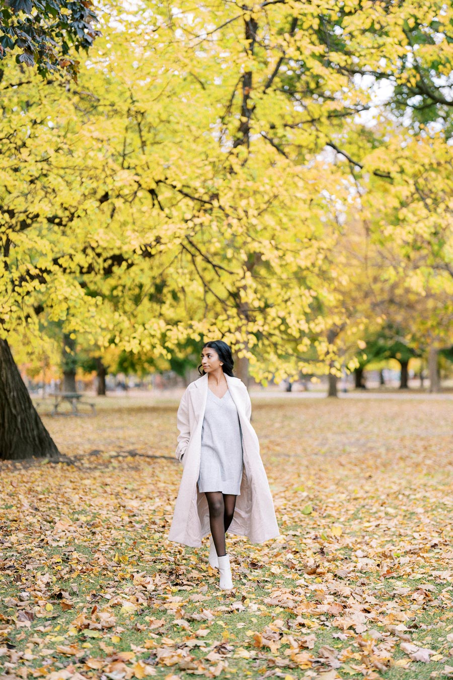 A person in a stylish outfit, wearing a light coat and dress, walks through an autumn park with vibrant yellow leaves and fallen foliage, capturing the essence of fall fashion and nature.