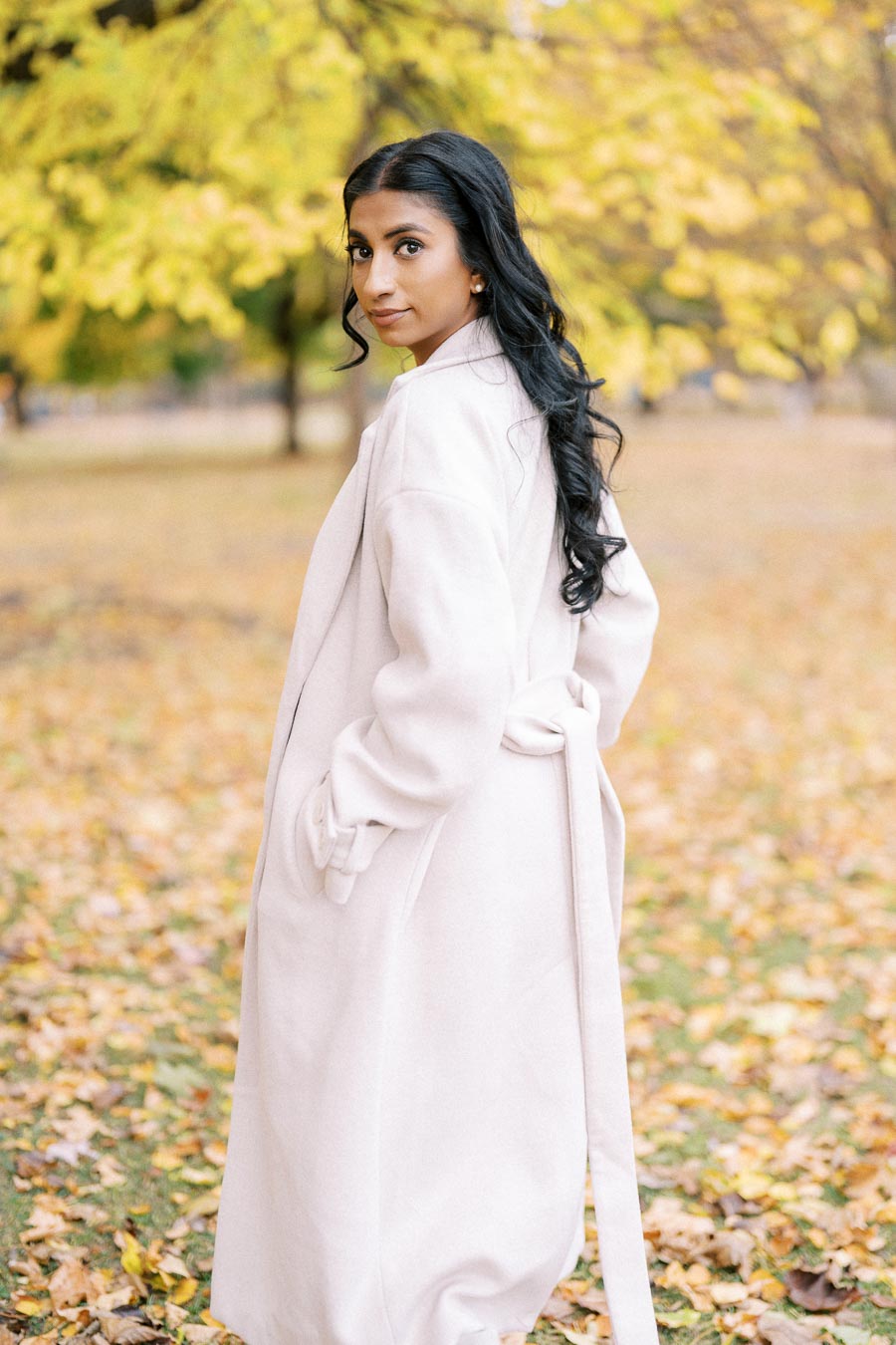 Elegant woman in a light pink coat glancing back in a sunlit park with vibrant yellow autumn leaves covering the ground.