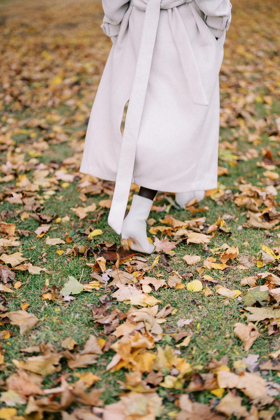 Person walking through autumn leaves on grass wearing a long beige coat and white ankle boots.