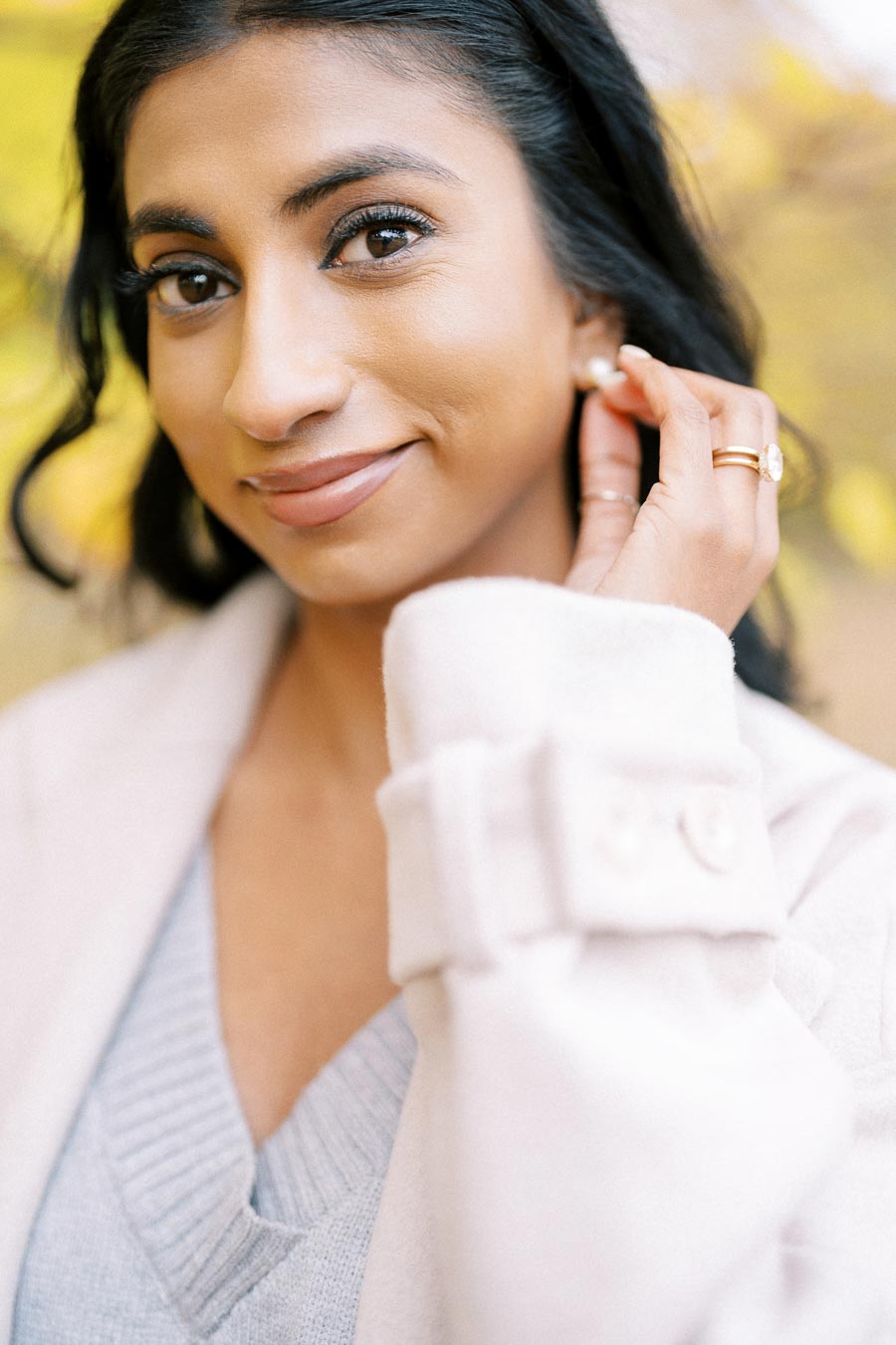 A woman smiling softly, wearing a light-colored coat and a v-neck sweater, with hand gently touching her ear, showcasing gold rings. The background features soft, blurred natural greenery, adding to the serene atmosphere.