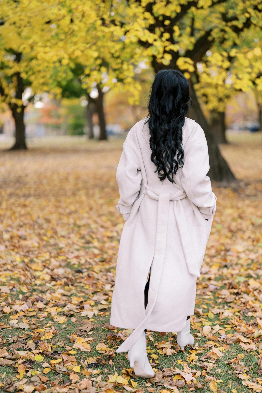 A woman in a long beige coat walks through a park covered with autumn leaves, surrounded by trees with yellow foliage.