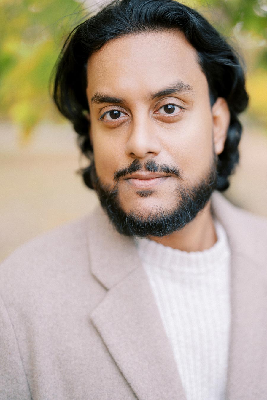 A person with a beard and black hair wearing a light-colored coat and sweater, standing outdoors with blurred greenery in the background.