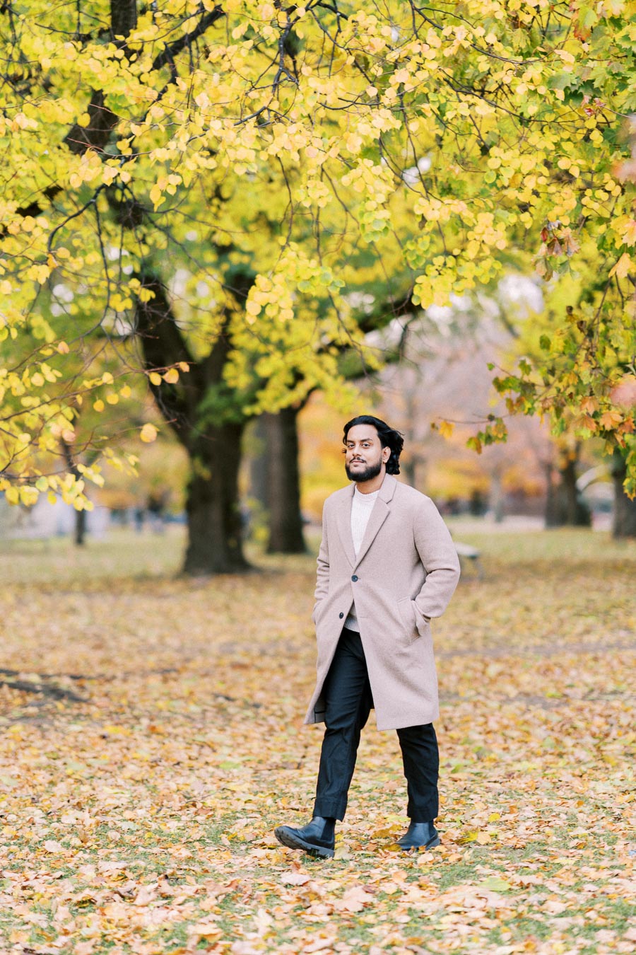 A man in a beige coat walking through a park covered in autumn leaves, with vibrant yellow-green trees in the background.