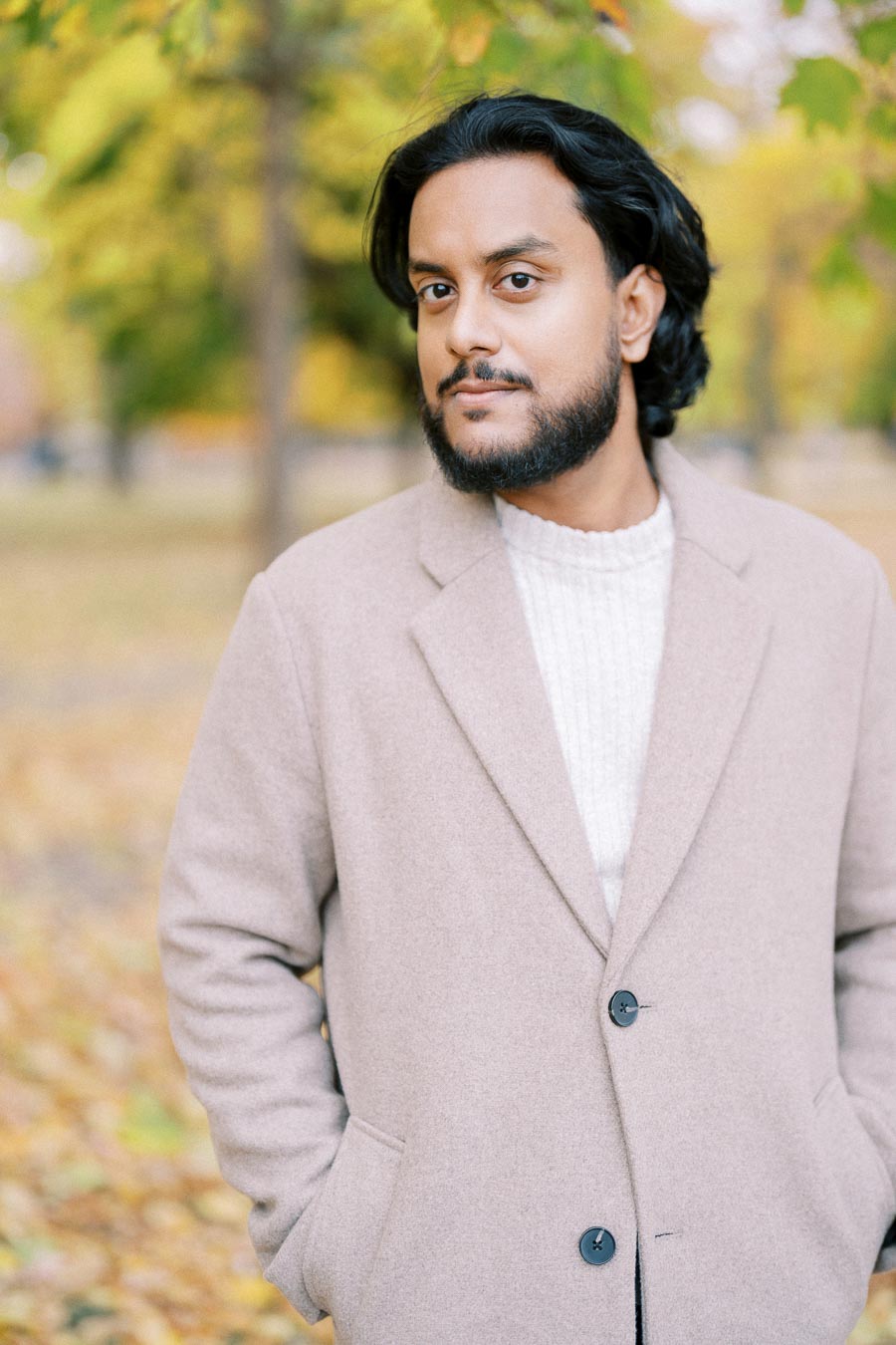 A person wearing a beige coat stands in a park during autumn, with yellow and green leaves visible in the background.