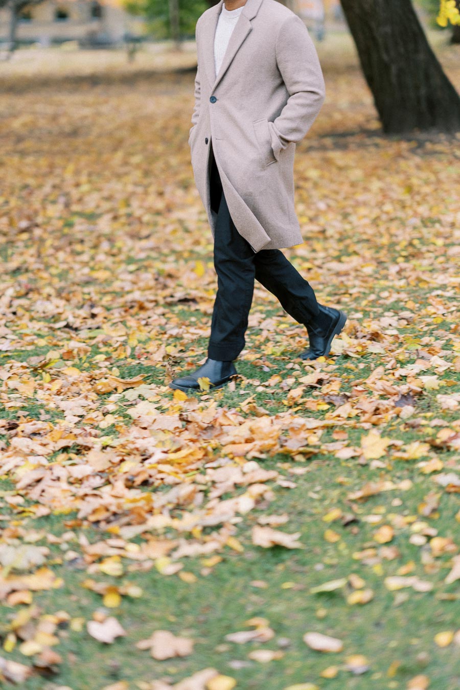 A person in a beige overcoat walks through a park covered with autumn leaves.