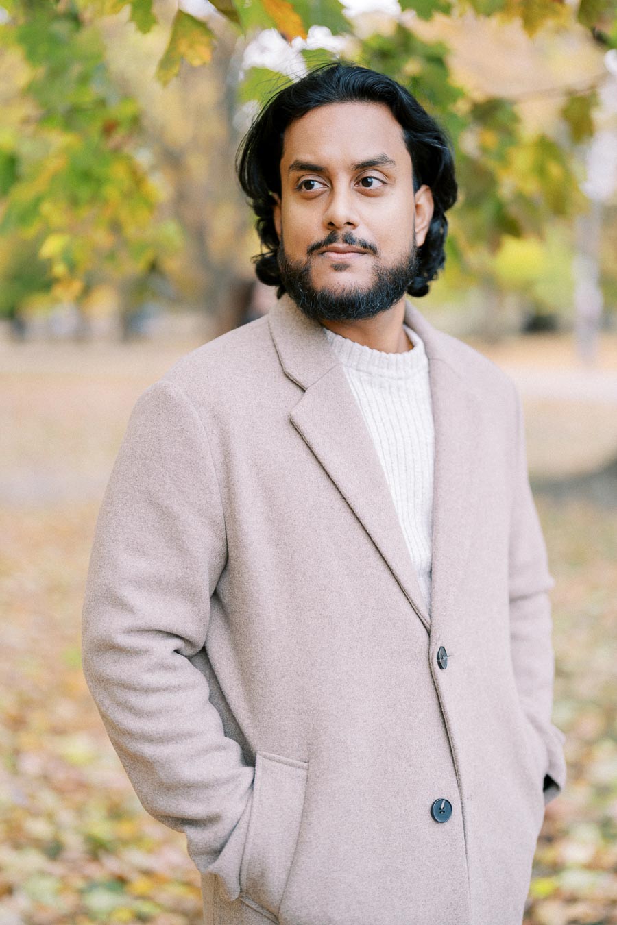 A man in a beige coat standing outdoors in a park during autumn, surrounded by colorful leaves.