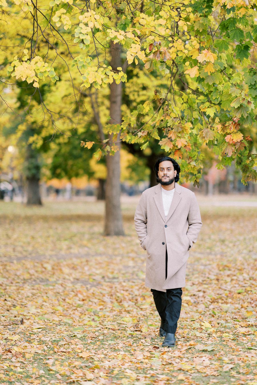 Man walking through a park on an autumn day with colorful fall foliage and fallen leaves, wearing a beige coat.