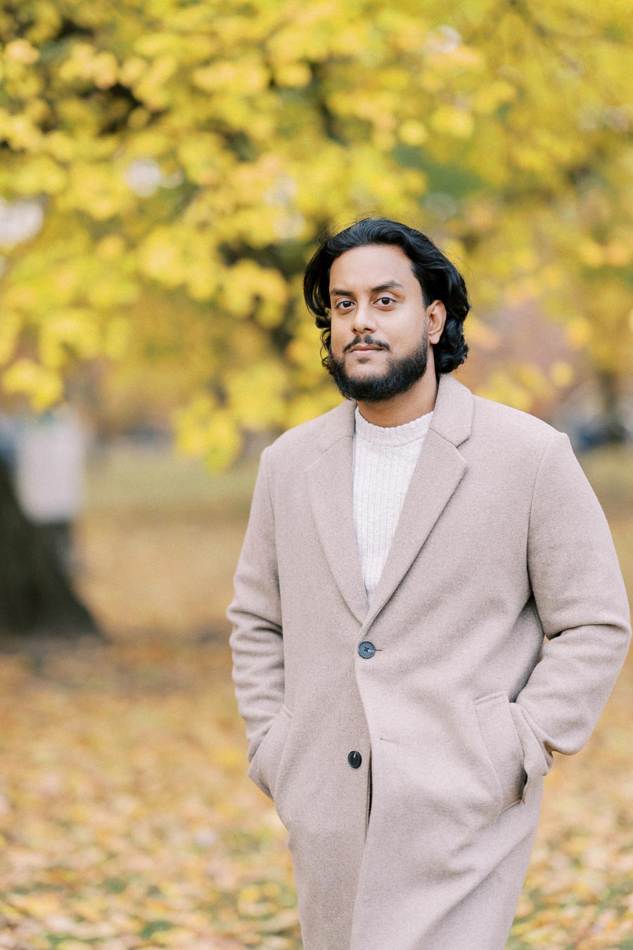 A person with dark hair and a beard wearing a beige overcoat stands in a park with vibrant yellow autumn leaves in the background, hands in pockets.