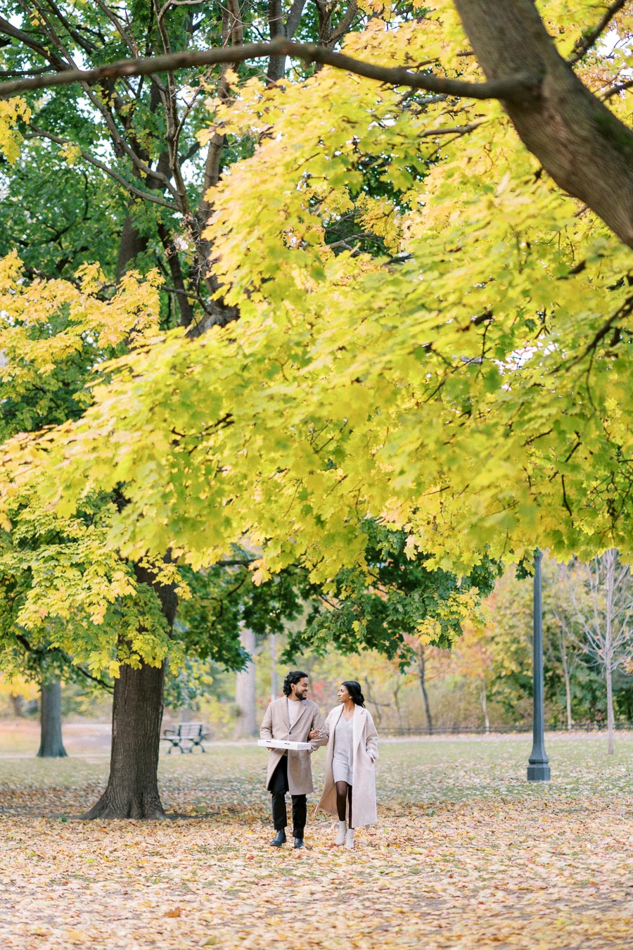 A couple walks arm in arm through a picturesque park during autumn, surrounded by vibrant yellow and green leaves, under a large tree with a lamp post nearby.