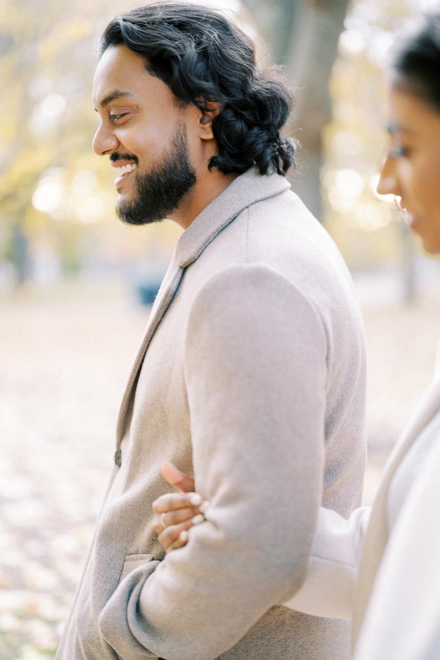 A man in a light-colored coat smiling while walking arm-in-arm with a woman on a park path during autumn.