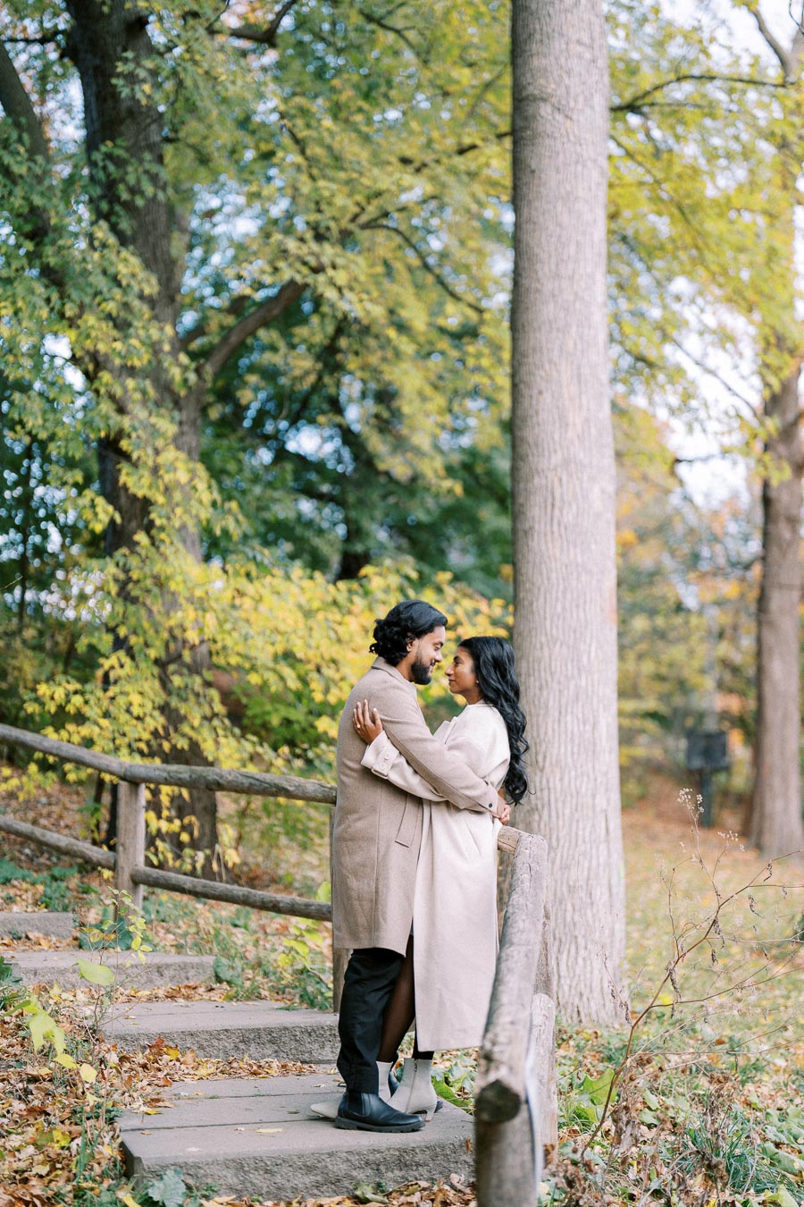 A couple embraces on a forest trail surrounded by autumn foliage, wearing long coats, conveying a romantic outdoor moment.