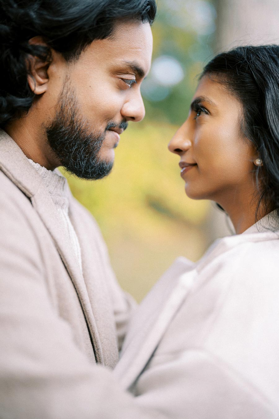 A couple gazes into each other's eyes, both wearing beige coats, set against a blurred natural background.