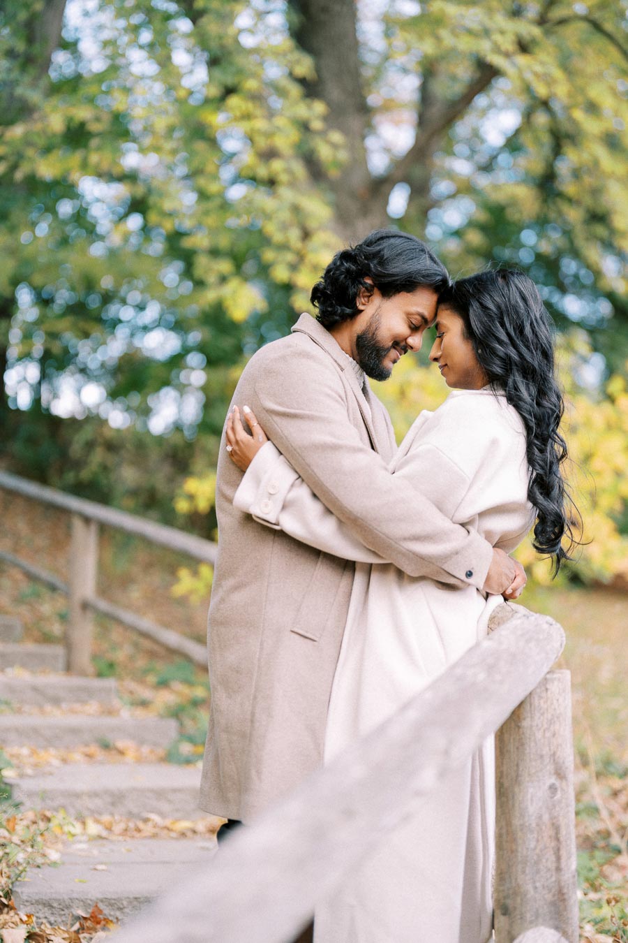 A couple in warm coats embraces on a scenic outdoor path with autumn leaves and green trees in the background, conveying a feeling of love and tranquility.