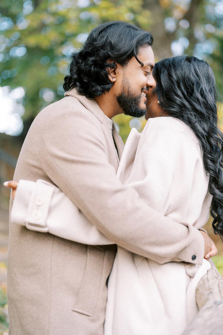 A romantic couple embracing outdoors in soft beige coats, sharing an intimate moment surrounded by blurred autumn trees.