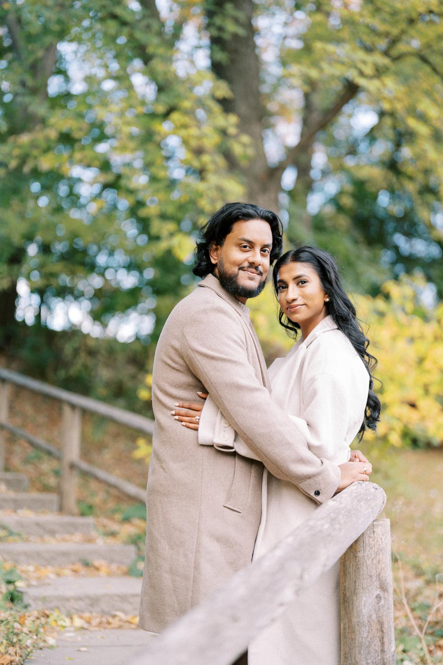 A couple in beige coats embrace on an outdoor wooden path surrounded by lush green and yellow trees, exuding warmth and natural beauty.