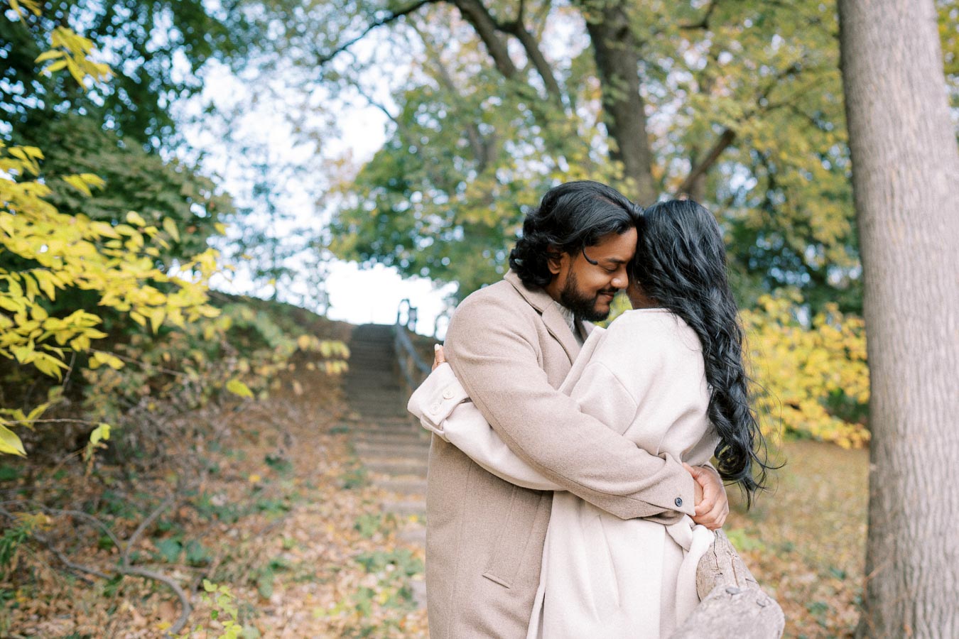 Romantic couple embracing in a park during autumn, with colorful fall foliage and a pathway in the background.