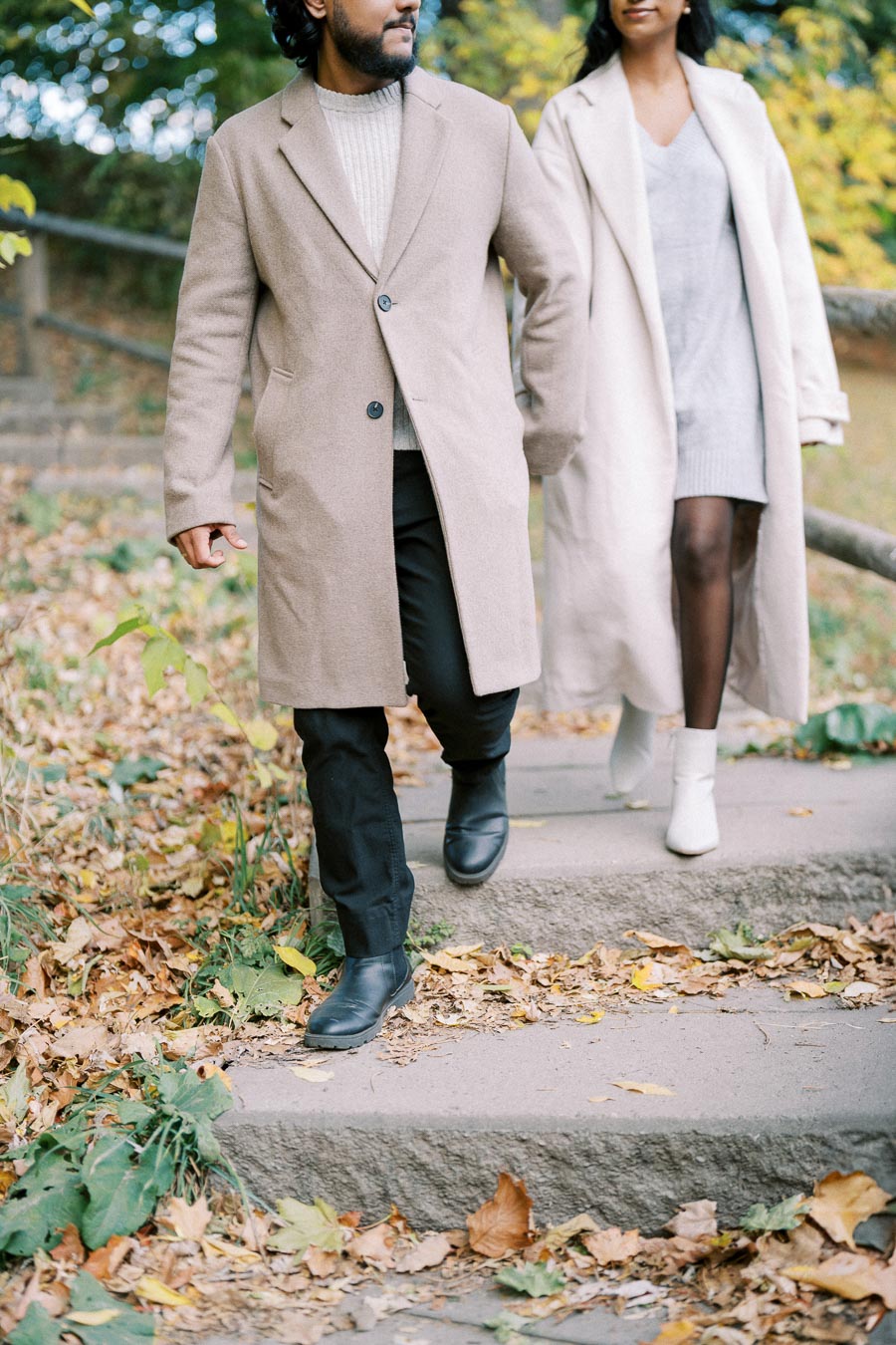 A stylish couple walking down outdoor stairs in autumn, wearing neutral-toned coats and surrounded by fallen leaves.