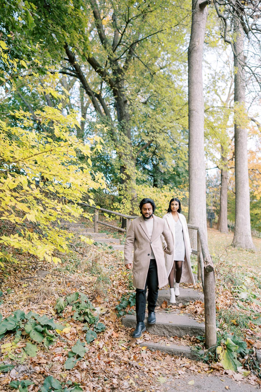 A couple walking down stone steps in a lush, green park during autumn, surrounded by trees with vibrant yellow and green leaves, both wearing stylish beige coats.