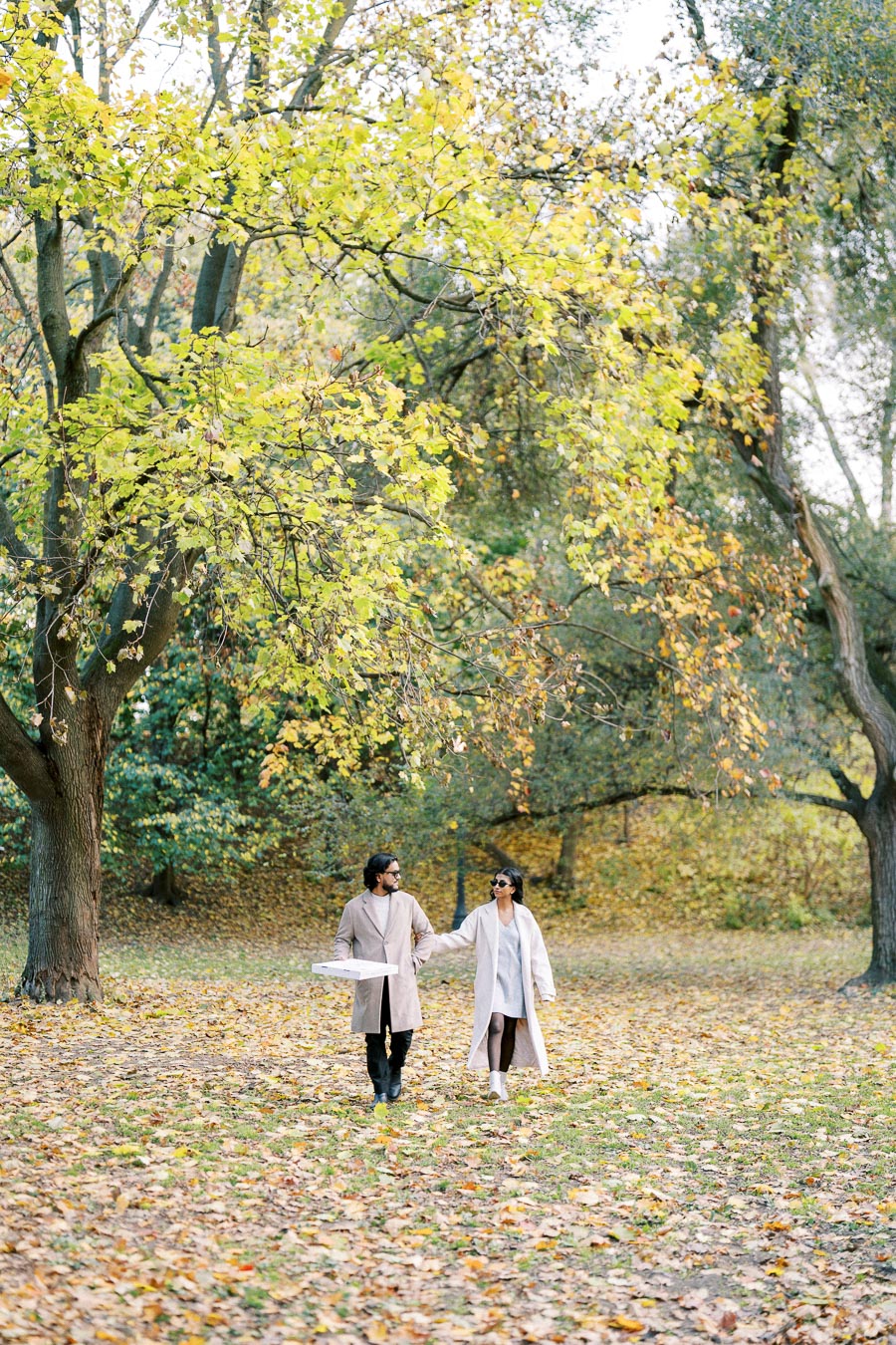 Couple walking through a park in autumn, surrounded by trees with vibrant yellow and green leaves, holding hands and a pizza box.