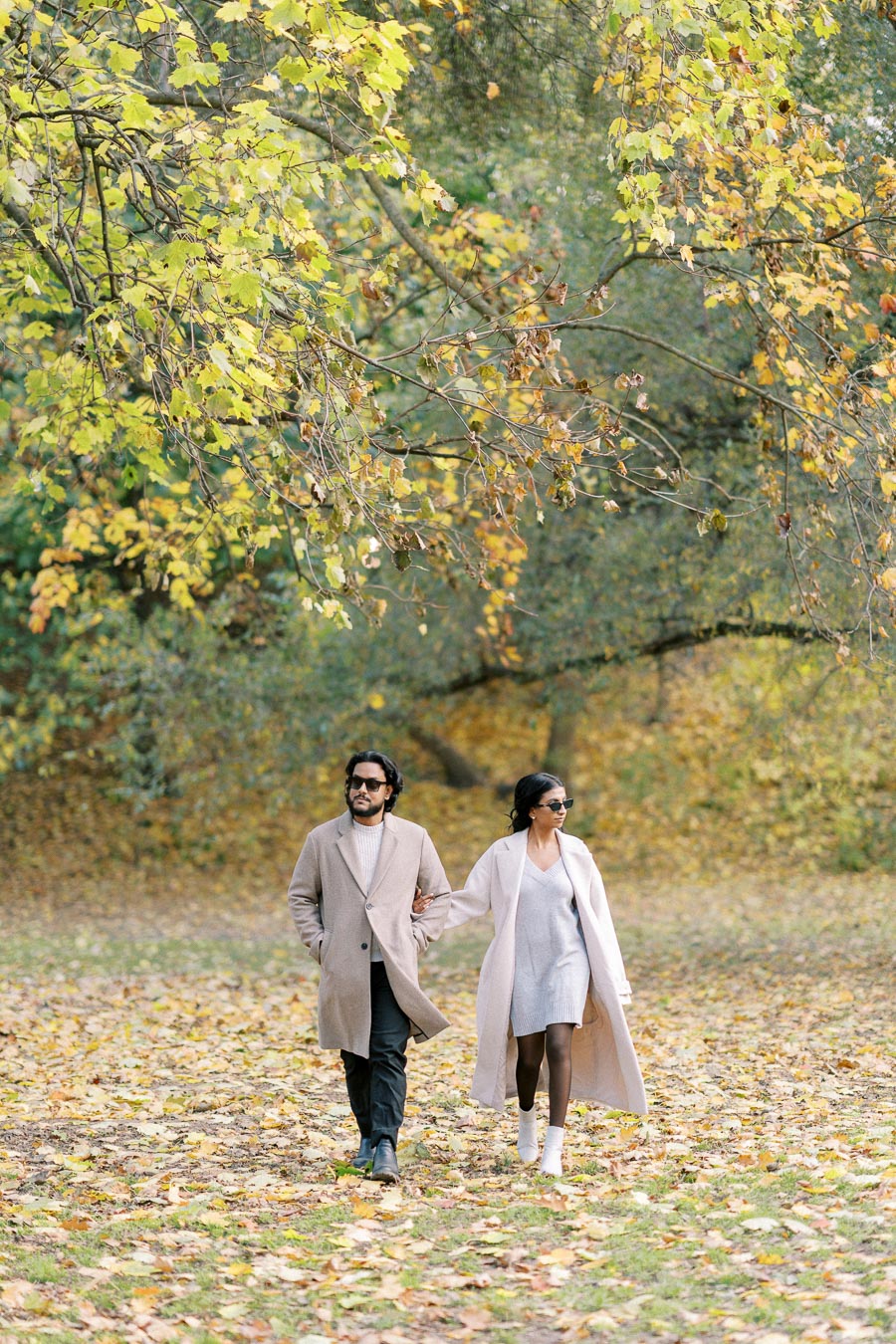 A stylish couple walking arm-in-arm through an autumn park, surrounded by colorful fall leaves and greenery, both wearing fashionable long coats and sunglasses.