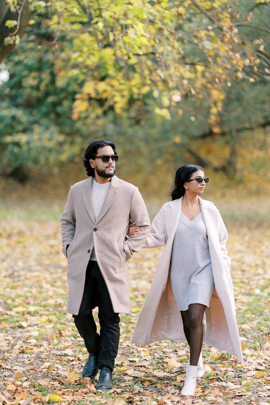 A stylish couple walking through a scenic autumn park, wearing fashionable beige coats and sunglasses, surrounded by fallen leaves and colorful foliage.