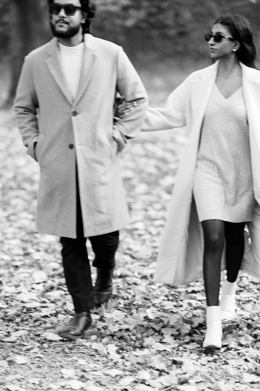 Black and white image of a stylish couple walking in an autumn park, wearing long coats and sunglasses, with fallen leaves on the ground.