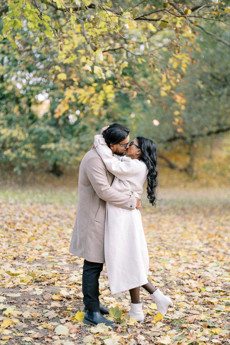 A couple embracing and kissing in a park during autumn, surrounded by fallen leaves and colorful trees.