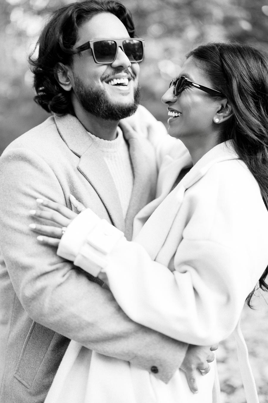 Black and white photo of a joyful couple in sunglasses laughing together, dressed in stylish coats, displaying a joyful moment outdoors.