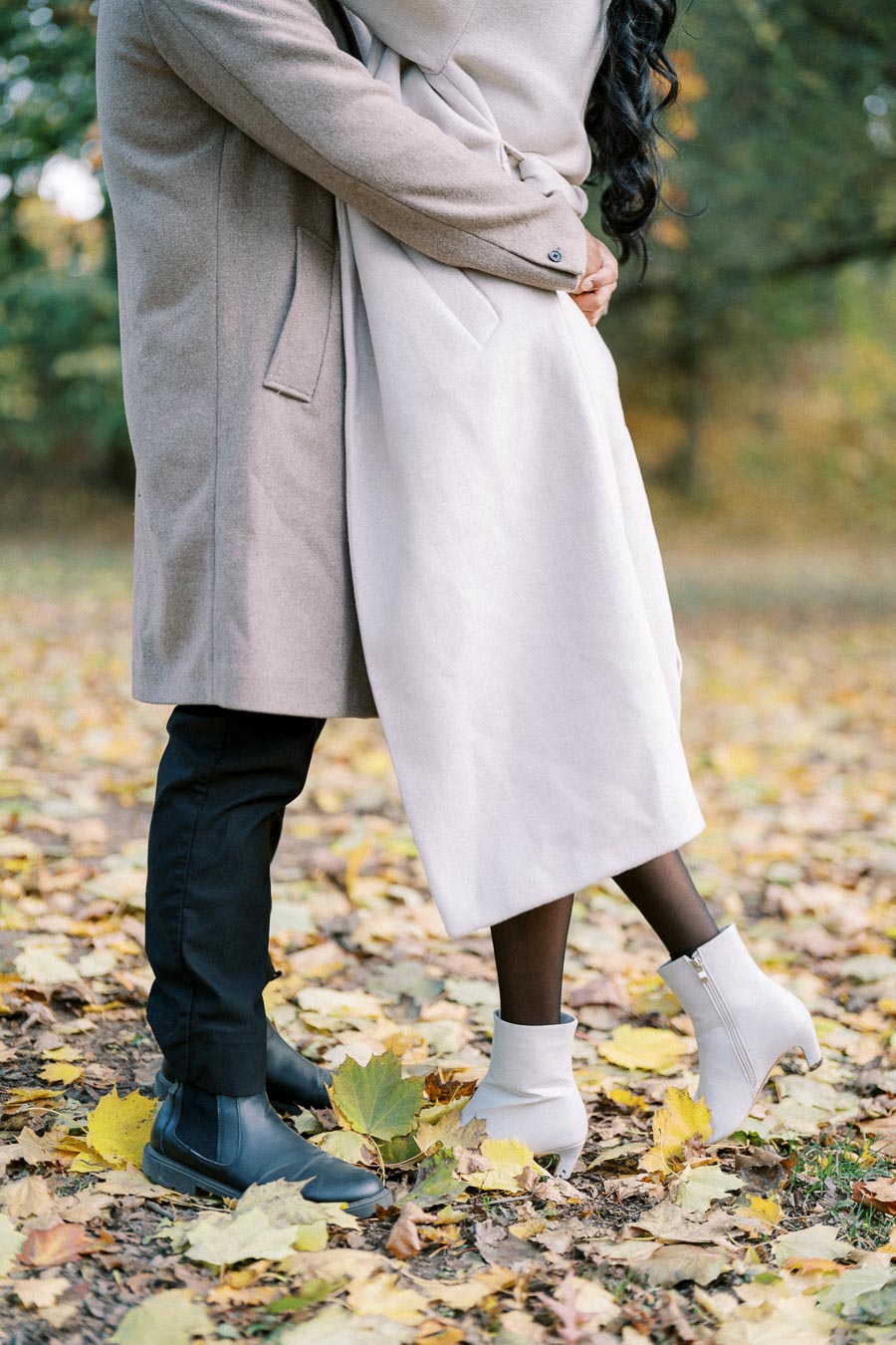 A couple embracing in a park with fallen autumn leaves, wearing stylish coats and ankle boots.