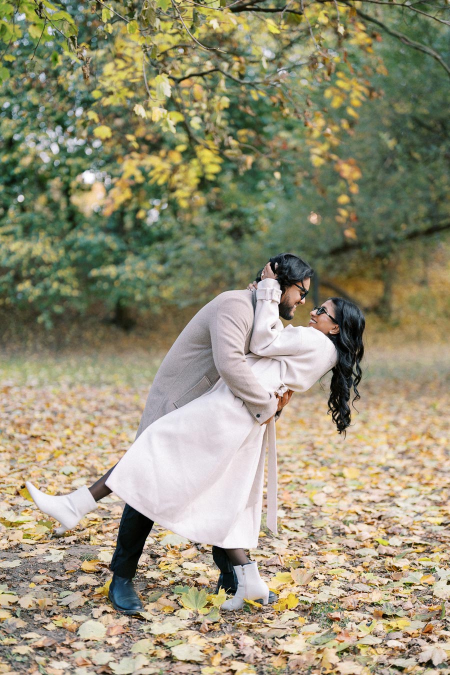 A couple in stylish autumn attire shares a romantic moment in a leafy park, with the man playfully dipping the woman amid colorful fallen leaves and vibrant foliage.
