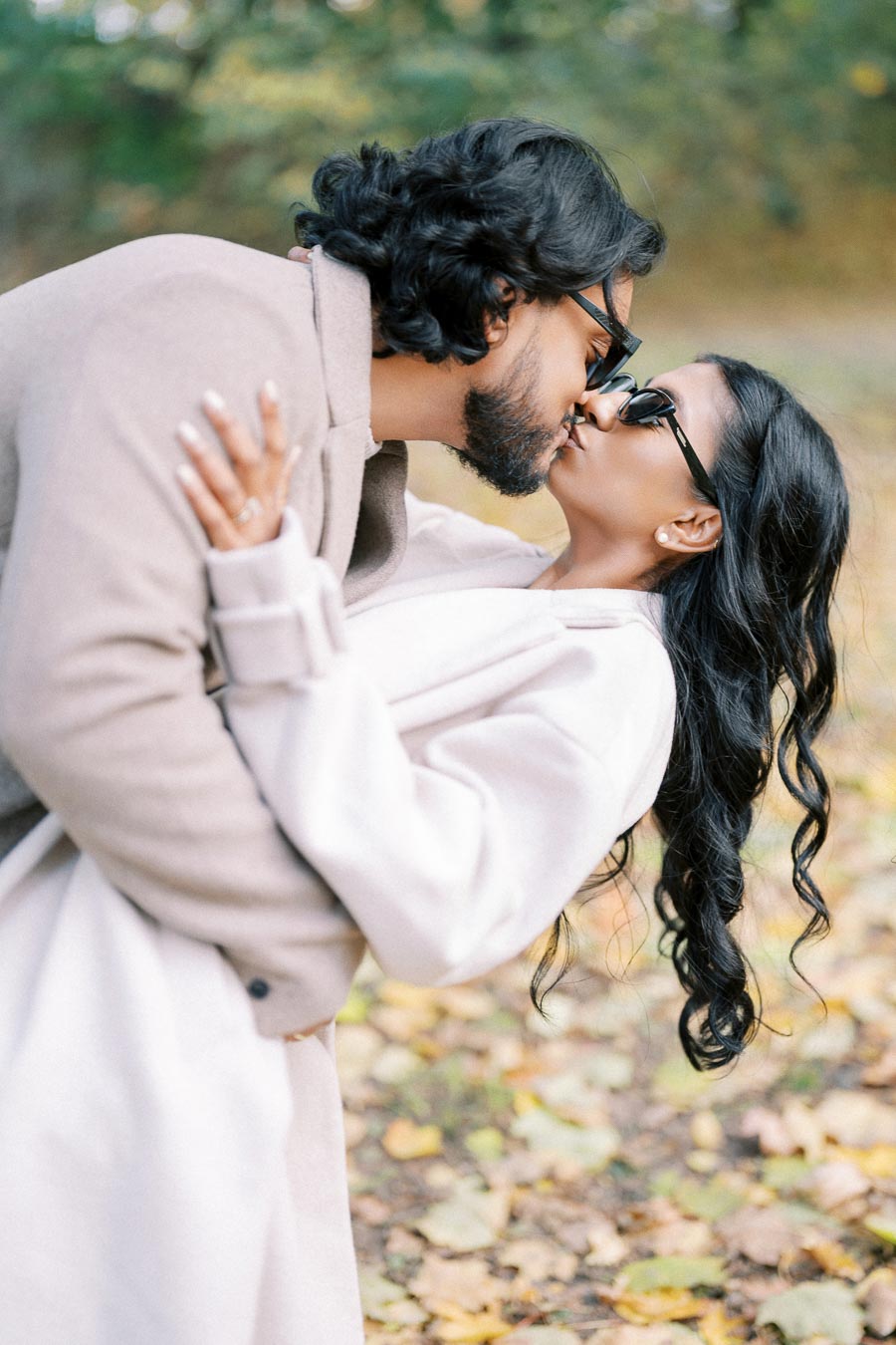 A couple kissing passionately outdoors, both wearing stylish coats and sunglasses, surrounded by a scenic background of autumn leaves and trees.